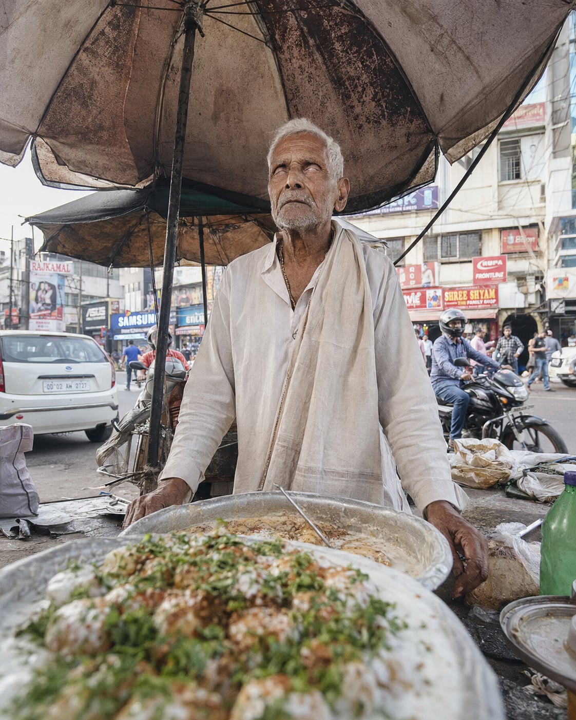 A white-haired elderly vendor with clouded, unfocused eyes stands tall and erect in a cream kurta beneath a large weathered umbrella, behind big aluminium pans of dahibara dumplings garnished with coriander and red chilli; behind him a busy modern street with a parked car, a helmeted motorcyclist, pedestrians and shop hoardings.
