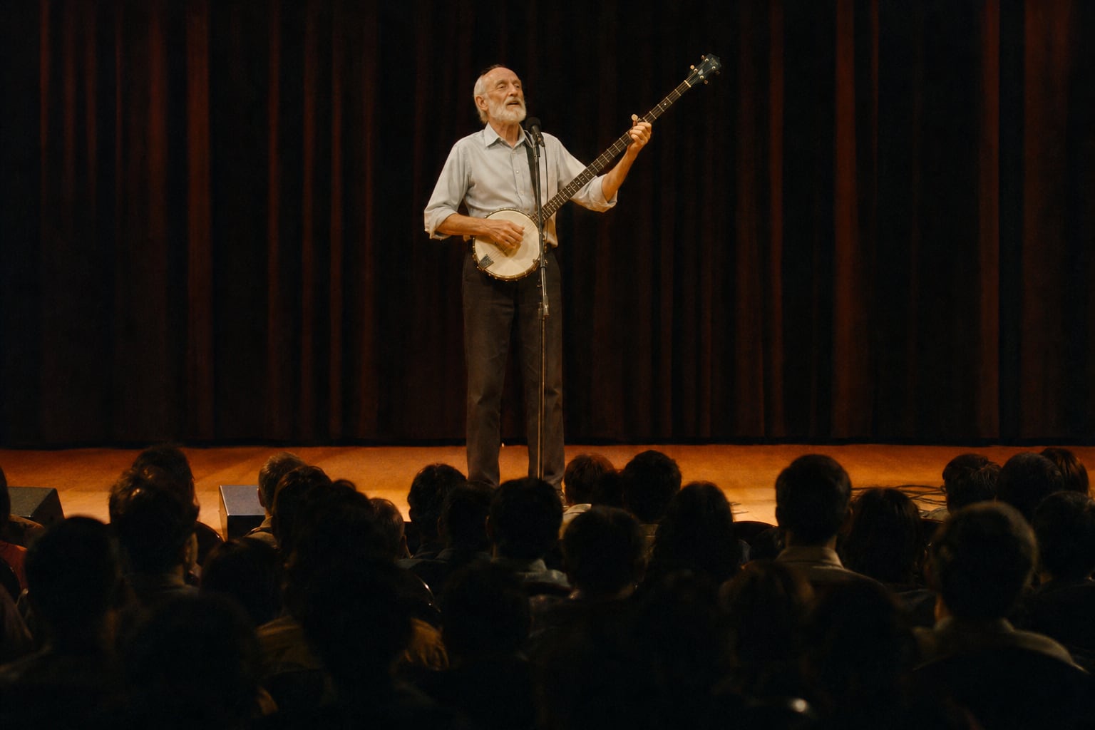 On a warmly lit auditorium stage, a tall, lean elderly man with a bald crown, white hair at the sides and a short white beard stands at a microphone playing a long-necked banjo, head tilted back mid-song; heavy curtains frame the stage, and in the foreground the dark silhouetted heads of a full Indian audience watch from the rows below.